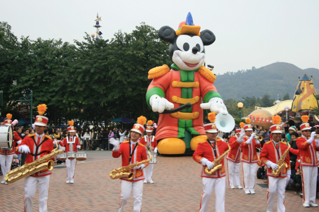 Gruppe von Menschen in Kostümen, die Instrumente spielen, vor einem Mickey-Maus-Floß während eines Umzugs, mit Bäumen, Gebäuden, Hügeln und einem klaren blauen Himmel im Hintergrund und einer Tafel mit Text auf der linken Seite.
