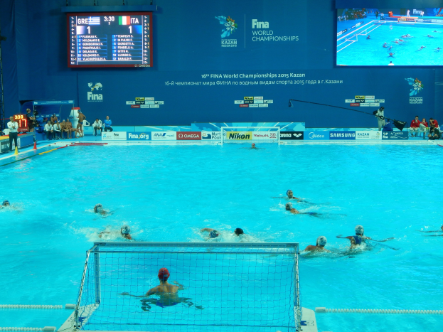 Eine Gruppe von Menschen spielt Wasserball in einem große Pool mit einem Netz in der Mitte, Zuschauern auf der linken Seite und einer Wand mit einem Bildschirm im Hintergrund bei den FINA-Weltmeisterschaften 2016 in Kazan.