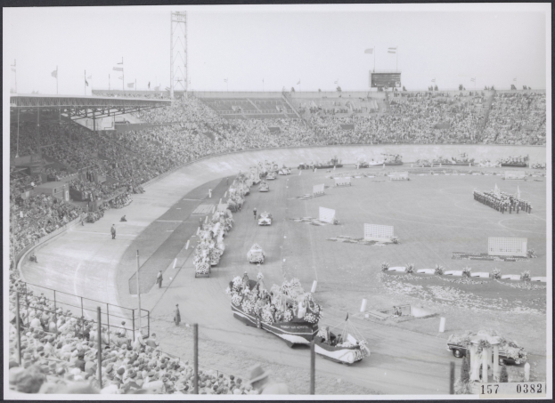 Schwarzes und weißes Foto einer Rennstrecke mit Zuschauern in einem Stadion, Fahrzeugen auf der Straße und Fahnen im Hintergrund.