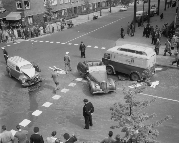 Eine Schwarz-Weiß-Szene eines Autounfalls an einer Straßenecke, mit mehreren Fahrzeugen, darunter einem Van, einer Gruppe von Menschen drumherum und im Hintergrund Gebäude, Bäume, Masten und Texttafeln.