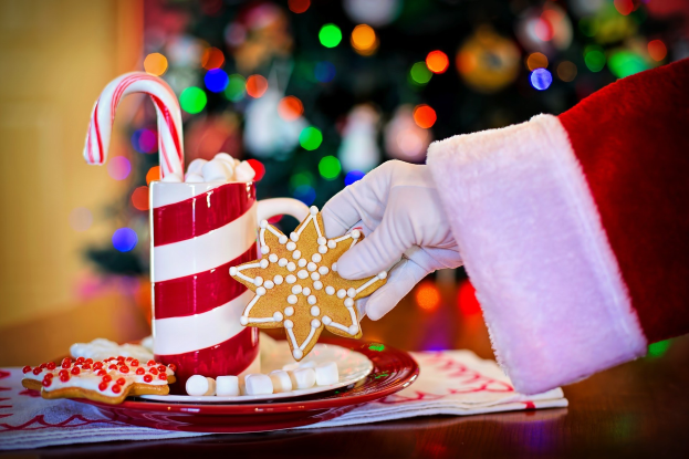 Ein Mensch mit einem Santa Claus Hut, der einen Becher heiße Schokolade und einen Zuckerstange hält und ein Teller mit Keksen und ein Tuch auf dem Tisch vor ihm stehen. Vor einem Weihnachtsbaum und einer Wand.