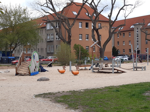 Spielplatz mit Rutsche, Schaukeln und anderem Equipment, umgeben von Gras, Bäumen, Gebäuden, Fahrzeugen und einem bewölkten Himmel.