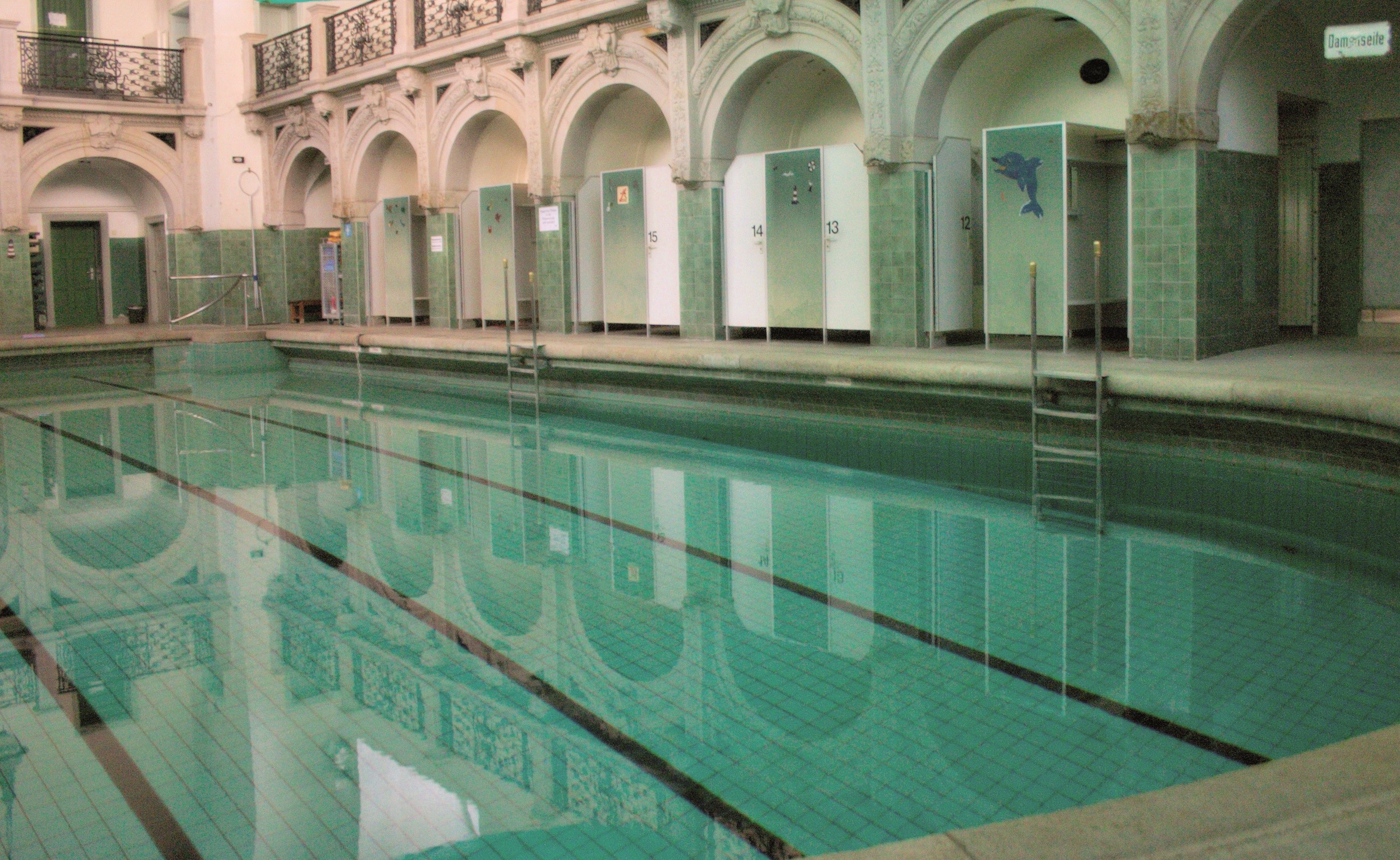 Innenbad mit klarem Wasser, umgeben von gefliestem Boden, Bogengängen, Pfeilern, Geländern, Türen und Texttafeln in einem großeen Gebäude.