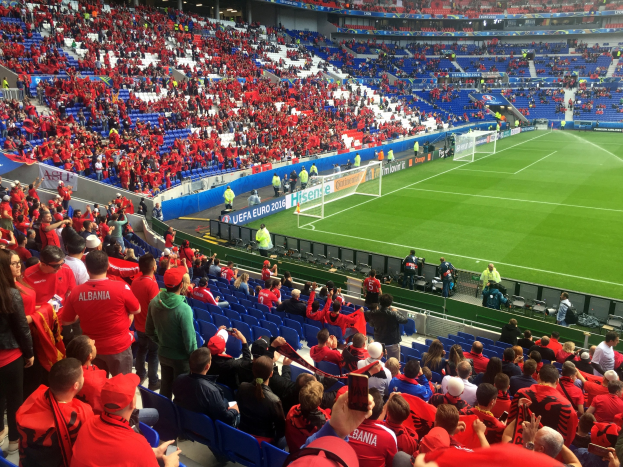Eine große Menschenmenge in einem Stadion schaut einem Fußballspiel zu, mit Sitzplätzen, Fahnen, einem Torpfosten und Deckenleuchten.