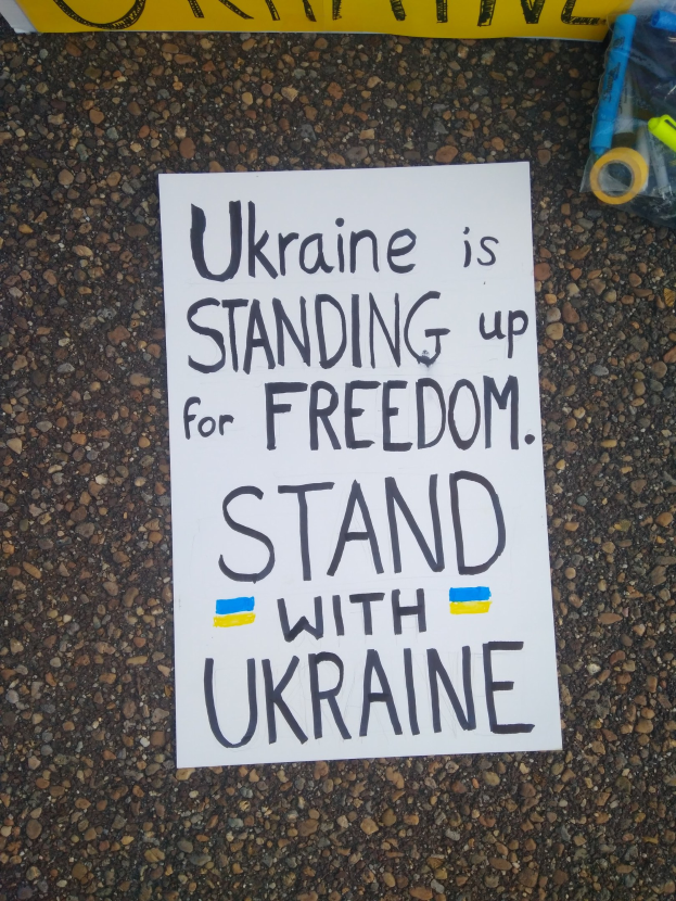 Weißer Papierbogen mit dem Text "Ukraine steht für Freiheit ein, steht mit Ukraine" auf dem Boden vor einer gelben Fahne, mit einer Plastikhülle mit Gegenständen auf der rechten Seite.
