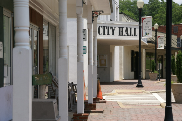 Rathausgebäude mit umliegender Infrastruktur, einschließlich Straßenlaternen, Schildern, Fahrzeugen, Verkehrskegeln, Stühlen, Topfpflanzen, Bäumen und einem bewölkten Himmel.