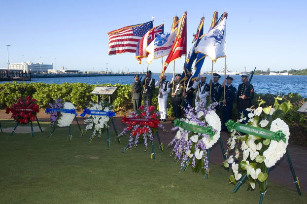 Eine Gruppe von Menschen vor einem Gewässer, mit Fahnen und Mützen, Blumensträußen auf dem Rasen und Schildern mit Text bei einer Gedenkfeier des National Park Service am Memorial Day.