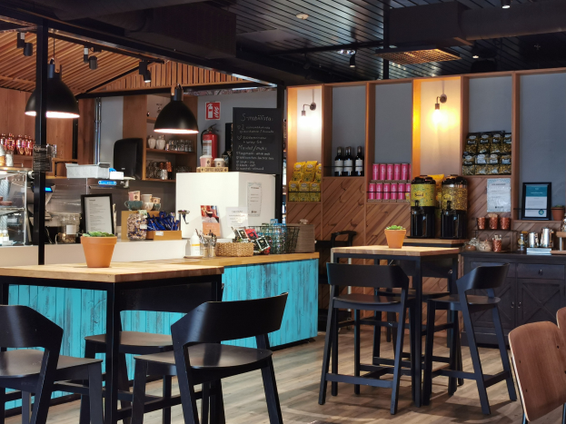 Inneneinrichtung eines Restaurants mit Holztischen, Stühlen, einer Topfpflanze auf einem Tisch, einer Schiefertafel, einem Kühlschrank, einem Feuerlöscher, Regalen mit Flaschen, Schränken mit Gegenständen, einem gerahmten Foto an der Wand, Deckenleuchten und Lampen.