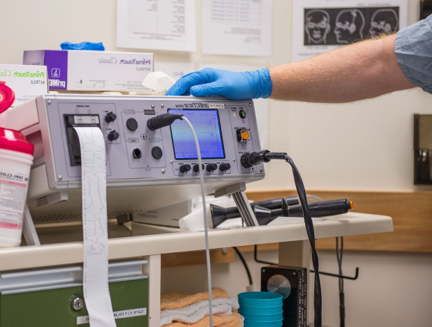 Eine Person in blauen Handschuhen arbeitet an einem medizinischen Gerät auf einem Tisch, der mit verschiedenen Gegenständen übersät ist, mit Papieren an der Wand im Hintergrund, was eine Krankenhausatmosphäre vermuten lässt.
