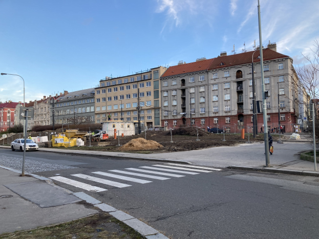 Baustelle für ein neues Wohnhaus im Vordergrund, mit parkenden Autos, Laternen, Bäumen und Gebäuden mit Fenstern, die eine Stadtstraße säumen, unter einem bewölkten Himmel.