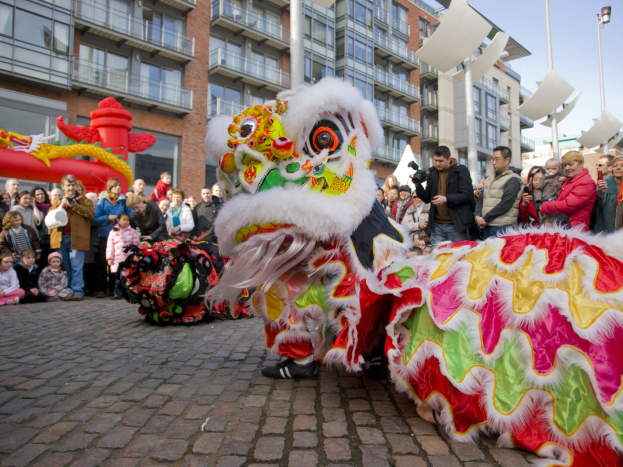 Ein lebendiges chinesisches Neujahrsfest in Amsterdam mit einem Löwen tanzen im Vordergrund und einer Menschenmenge mit Kameras in der Hand vor einem Gebäudehintergrund, Laternenmasten und einem klaren blauen Himmel.