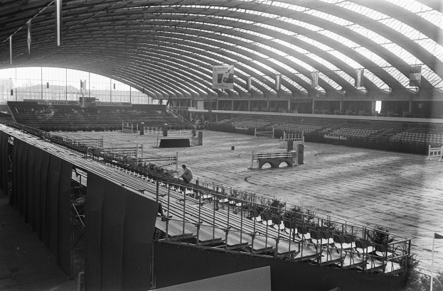 Schwarze und weiße Aufnahme eines großen Innenstadions mit einem Holzfußboden, einem Geländer, einigen Menschen, Stühlen, Bannern, Pfählen, Lampen und anderen Gegenständen, mit Gebäuden und Himmel im Hintergrund.