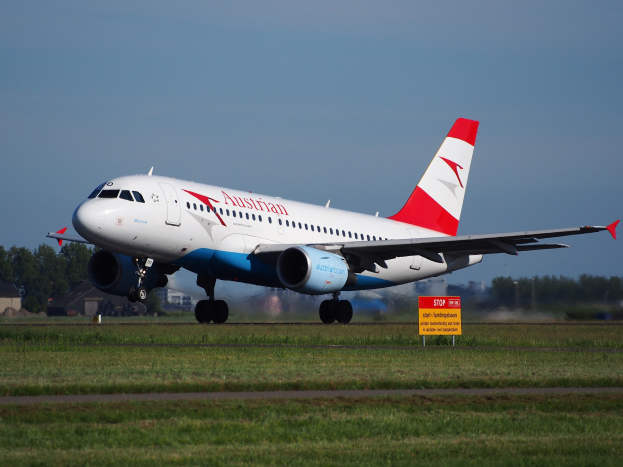 Austrian Airlines Airbus A320-200 startet von Frankfurt Airport mit einer Tafel im Vordergrund, Gras, Bäumen, Gebäuden und einem klaren blauen Himmel im Hintergrund.