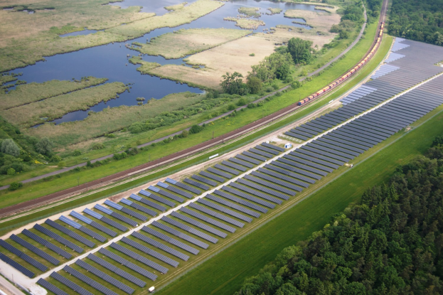 Luftaufnahme einer Solar-Farm mit Panelen in einem grasbewachsenen Feld, umgeben von Bäumen und Wasser, mit einem Zug auf einer nahen Schiene.