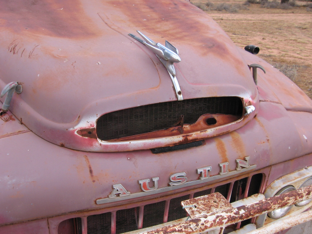 Alter rostiger Lkw auf einem Feld mit Bäumen und einem klaren blauen Himmel, der Spuren einer langfristigen Witterungseinwirkung zeigt.