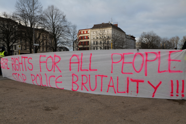 Eine Gruppe von Menschen hält ein Transparent mit der Aufschrift 'Rechte für alle Menschen Stoppt Polizeigewalt' auf dem Boden mit einem Straßenschild, einem Schild, Bäumen, Gebäuden mit Fenstern und einem bewölkten Himmel im Hintergrund.