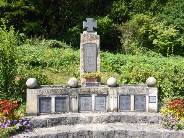 Ein Gedenkstein mit einem Kreuz darauf, umgeben von Pflanzen und Bäumen, mit eingravierten Wörtern und Zahlen, wahrscheinlich Kriegsgräber commemorating.