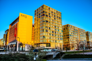 Eine belebte Stadtlandschaft mit hohen Geb├Ąuden, darunter das Jurys Inn Birmingham City Centre, mit Kraftfahrzeugen auf der Stra├če, Stra├čenmasten, Stra├čenlaternen, Verkehrsampeln, B├╝schen, B├Ąumen und einem klaren blauen Himmel.