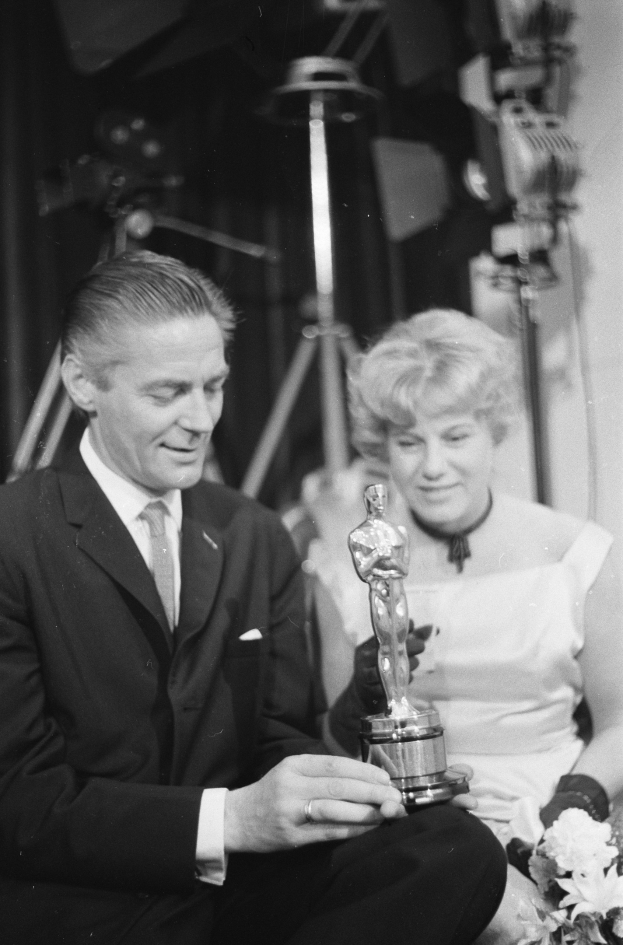 Ein Mann in einem schwarzen Blazer, einem weißen Hemd und einer Krawatte hölt eine Oscar-Statue, waehrend er neben einer Frau in Abendgarderobe sitzt, die einen Blumenstrau├č hölt, mit Hintergrundbeleuchtung in einem Schwarz-Wei├č-Bild.