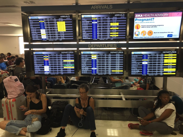 Menschen sitzen auf einem Flughafenboden, einige halten Handys, mit verstreuten Taschen und Bildschirmen, die Zika-Virus-Informationen anzeigen.