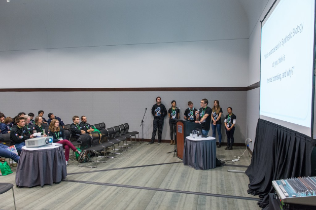 Eine Person, die in ein Mikrofon spricht, bei einer Besprechung mit sitzenden Zuschauern, Teammitgliedern mit Ausweisen, einem Präsentationsbildschirm und einem Videoabspieler in einem leeren Büroraum.