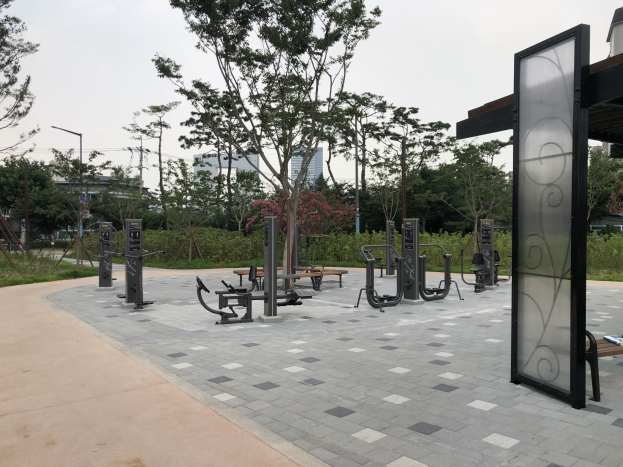 Outdoor-Fitnesspark mit Bänken, Bäumen, Pflanzen, Gras, Pfosten, Lichtern, Drähten und Gebäuden unter dem Himmel.