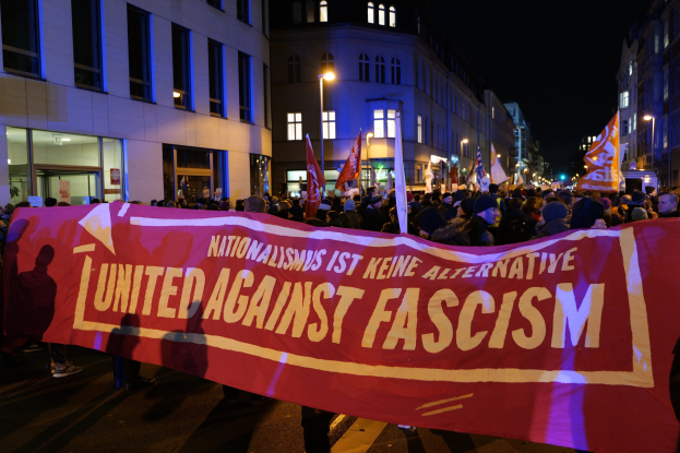 Eine Gruppe von Menschen marschiert auf einer Straße, die ein Banner mit der Aufschrift 'Einheit gegen Faschismus' trägt, mit Gebäuden, Laternen und Schildern an der Straße und dem Himmel im Hintergrund.