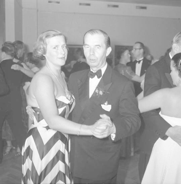 Ein Mann in einem schwarzen Blazer, weißem Hemd und schwarzen Hosen tanzt mit einer Frau in einem unbekannten Outfit auf einer formellen Veranstaltung und hält dabei Hände.