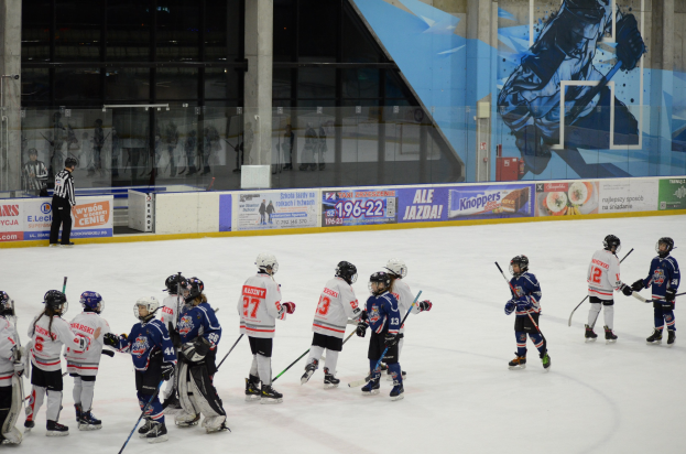 Young people in helmets and hockey gear playing ice hockey on an indoor rink with banners, pillars, and glass walls in the background.