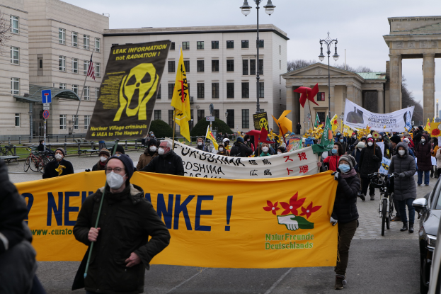 Eine große Gruppe von Menschen marschiert auf einer Straßendemonstration gegen Atomkraft in Deutschland, mit Schildern und Fahnen auf der rechten Seite und Fahrzeugen, Gebäuden, Bäumen und einem Tor im Hintergrund.
