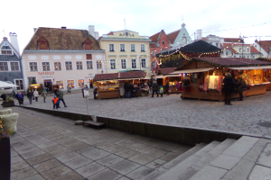 Ein lebendiger Weihnachtsmarkt in Tallinn, Estland, mit Menschen um geschmückte Stände, festlicher Beleuchtung und Gebäuden mit Fenstern im Hintergrund, unter einem bewölkten Himmel, mit Treppen, die zum Markt führen und Töpfen mit Pflanzen, die herumstehen.