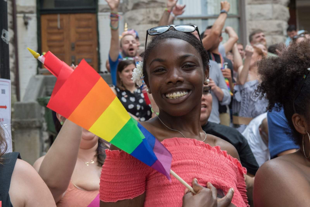 Eine Gruppe von Menschen vor einem Gebäude stehend, wobei eine Frau in der Mitte eine Regenbogenfahne hält; das Gebäude hat Fenster und eine Tür und ein Schild mit Text ist sichtbar.