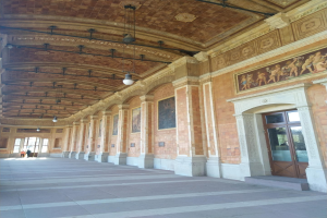 Langer Flur mit gefliestem Boden, Säulen, Türen, Wandrahmen, Deckenleuchten und mehreren Personen, der einem belebten Rathausinnenraum ähnelt.