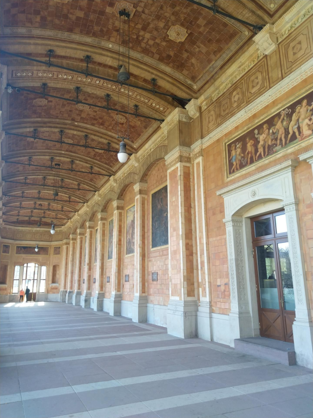 Langer Flur mit gefliestem Boden, Säulen, Türen, Wandrahmen, Deckenleuchten und mehreren Personen, der einem belebten Rathausinnenraum ähnelt.