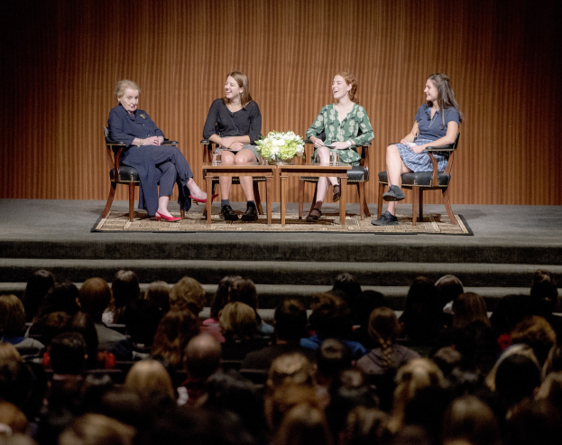 Vier Frauen auf Stühlen auf einer Bühne vor einem Publikum sitzend, mit einem Tisch mit einer Vase und anderen Gegenständen dahinter und Menschen auf einer Treppe darunter.