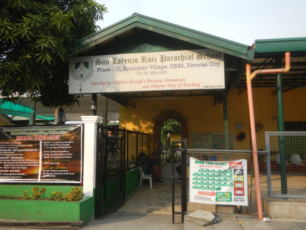 Der Eingang der San Lorenzo Ruiz Pfarrschule zeigt ein Schild mit Text, einen Metallzaun, Menschen auf Stühlen unter einem Dach, Pfosten, ein Schild, Pflanzen, einen Baum und einen bewölkten Himmel.