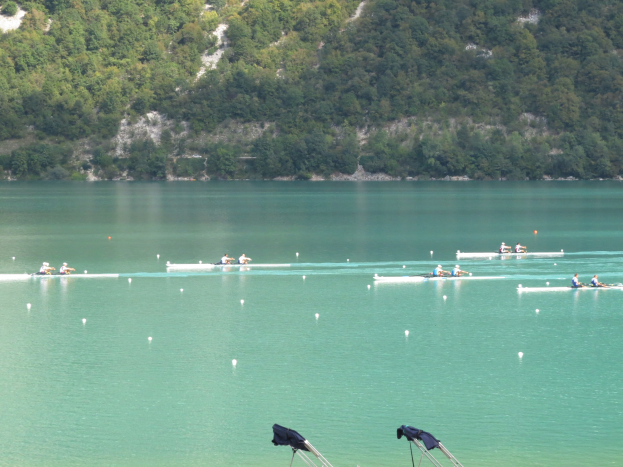 Gruppe von Menschen bei einem Ruderwettbewerb auf einem Gewässer mit Bäumen und einem Hügel im Hintergrund, Kleidung auf Ständen darunter.