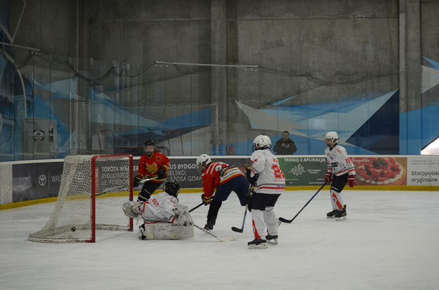 Eishockeyspieler in Helmen und Uniformen konkurrieren in der Nähe eines Torpfostens auf einer Indoor-Eisbahn, mit Bannern und einer Wand im Hintergrund.