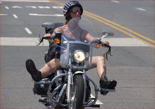 Professionelle Radrennfahrerin Katie Archibald trägt einen Helm, eine Schutzbrille, Handschuhe und Schuhe und fährt ein Fahrrad eine Straße entlang neben einer Menge Menschen. Im Hintergrund gibt es ein Banner mit Text, eine Wand, Bäume und ein Gebäude.