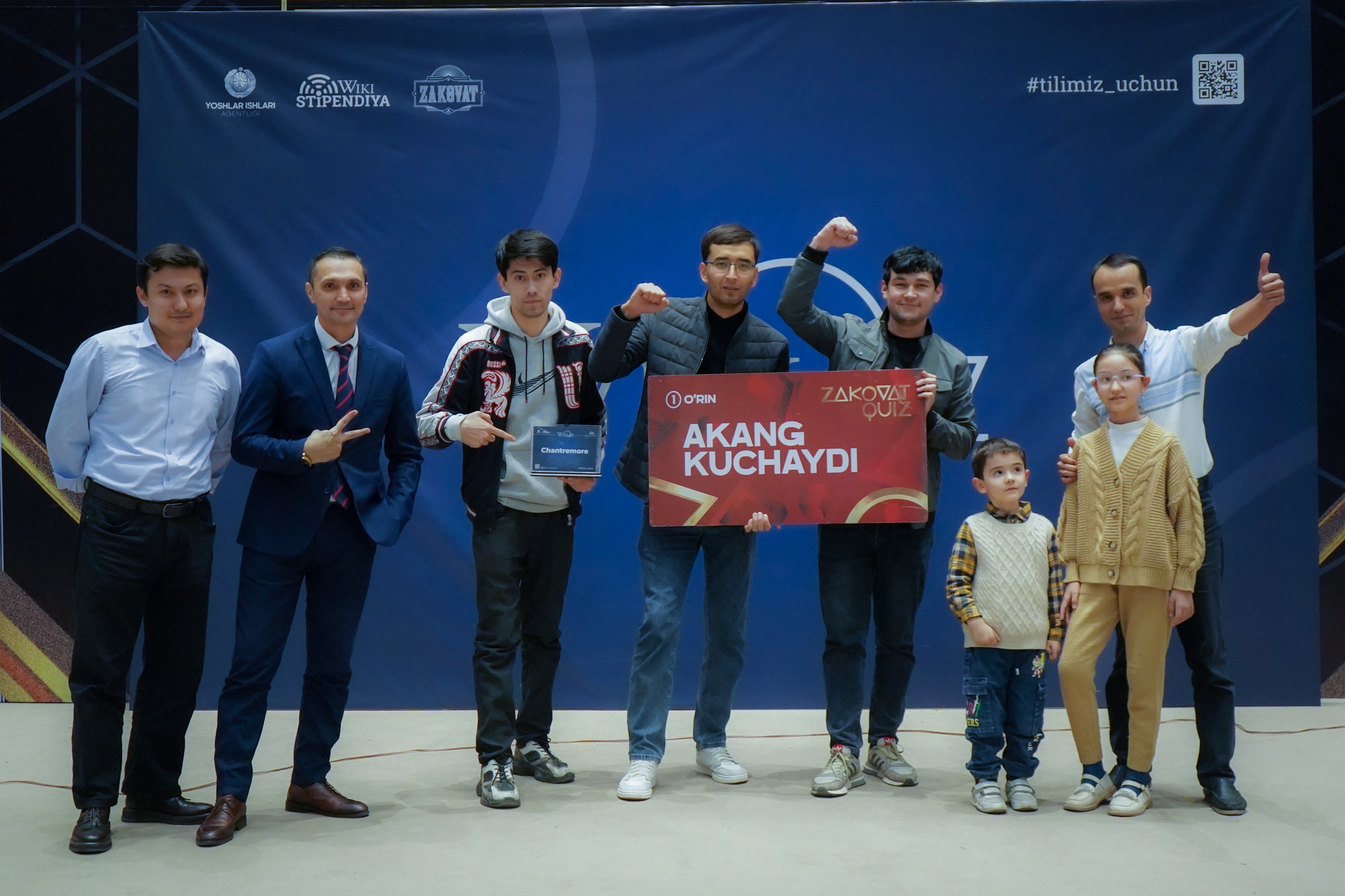 Gruppe von Menschen auf einer Bühne, die eine Tafel mit dem Text 'Akang Kuchaydi - Zakovat Quiz' halten, mit einem Banner und einem Strichcode im Hintergrund.