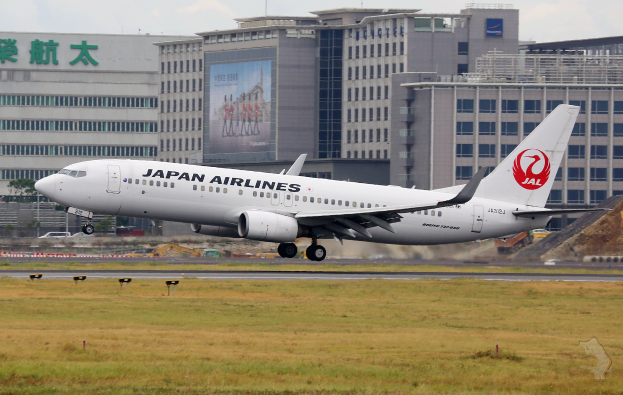Japan Airlines Airbus A320-200 beim Starten vom Tokyo Narita International Airport, umgeben von grünem Gras mit im Hintergrund sichtbaren Geb├Ąuden und Fahrzeugen unter einem teilweise bew├Âlktem Himmel.