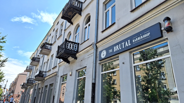 Brutalistischer Barbershop-Laden in Berlin mit Betongebäuden, Fenstern, Geländern, Balkonen und Schildern, umgeben von Bäumen, Laternenmasten und Fußgängern unter einem bewölkten Himmel.
