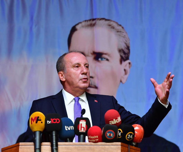 Ein Mann im Anzug steht an einem Podium mit Mikrofonen und spricht hinter einem Bildschirm, als ob er eine Ankündigung des Rücktritts von türkischem Präsident Recep Tayyip Erdogan kommentiert.