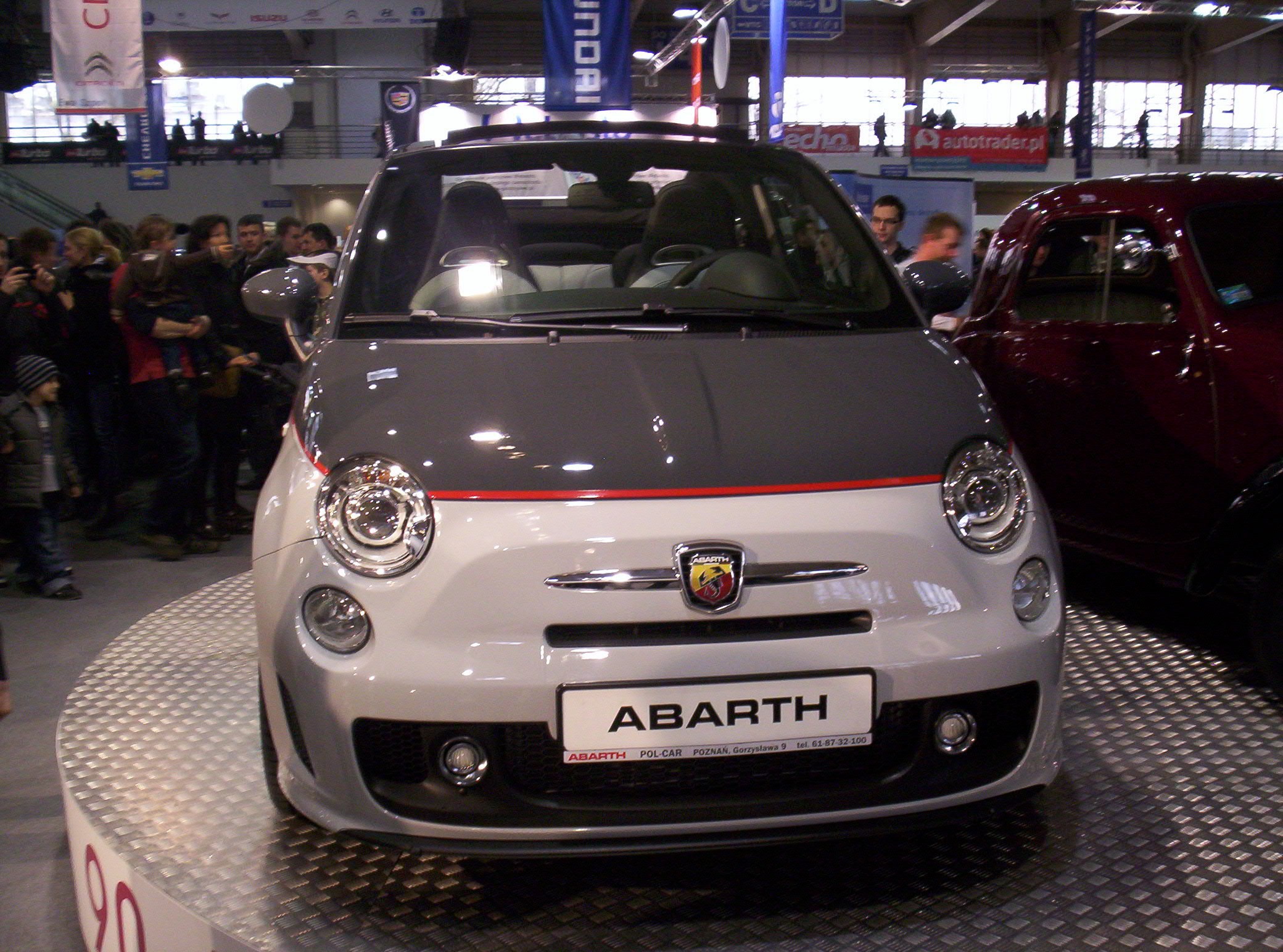 Ein roter und schwarzer Fiat 500 Abarth auf einer Autauschstellung, umgeben von Menschen und Bannern im Hintergrund.