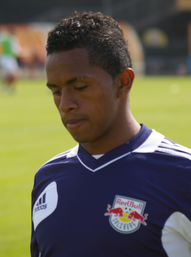 Ein junger Mann in einem blauen T-Shirt steht auf einem Fussballfeld, er wirkt wie ein Flügelspieler mit kurzen dunklen Haaren und einem entschlossenen Gesichtsausdruck vor einem unscharfen Hintergrund.