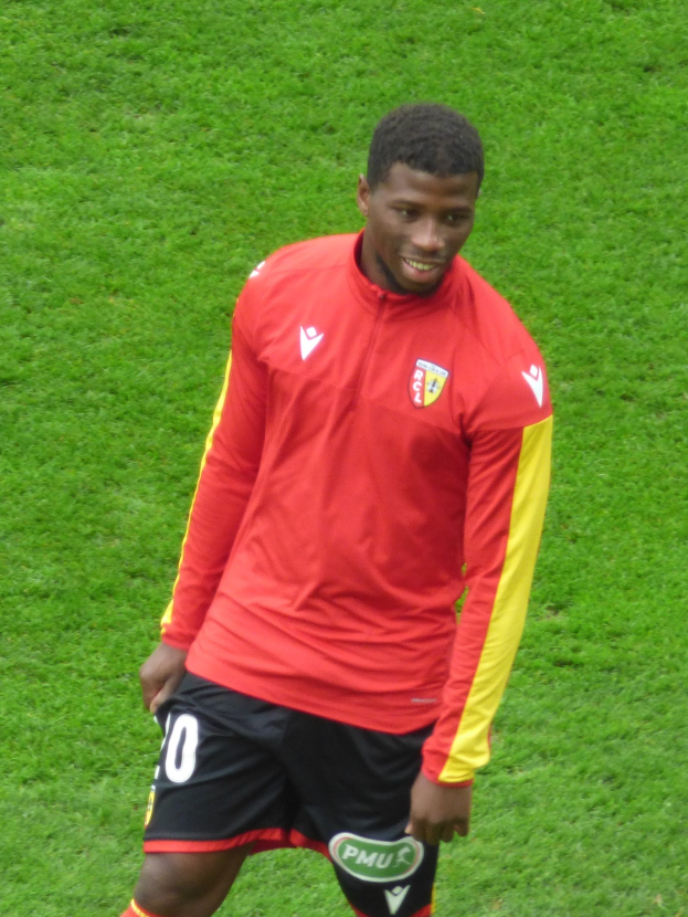 Ein Mann in rotem T-Shirt und schwarzen Shorts steht lachend auf einem Fußballfeld, positioniert als Flügelspieler.