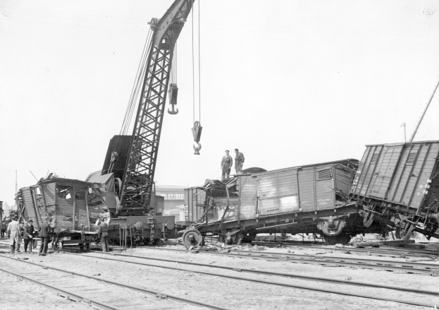 Schwarzes und weißes Bild eines Zugunglücks mit einem Kran, der einen Zugwagen auf die Schienen hebt, mehrere Menschen in der Nähe, Gebäude und ein klarer Himmel im Hintergrund.