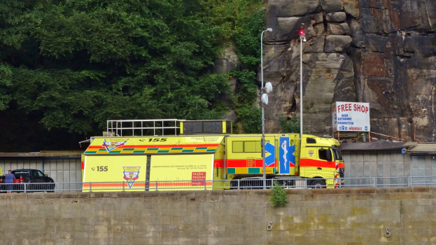 Ambulanz auf der Seite einer Straße neben einer Klippe geparkt, mit verschiedenen Objekten wie einer Wand, einem Geländer, Pfosten, Lampen und Texttafeln im Vordergrund und Bäumen und einem Auto im Hintergrund.