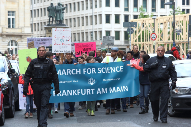 Eine Gruppe von Menschen marschiert auf einer Straße und hält ein "March for Science Frankfurt am Main"-Schild hoch, während Autos neben ihnen fahren und Gebäude, Statuen und Bäume im Hintergrund zu sehen sind.