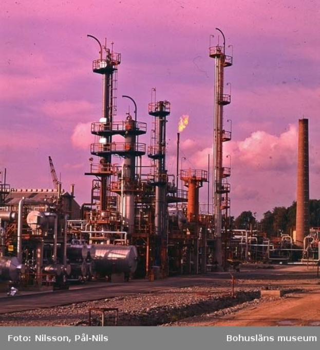 Ölraffinerie mit ausgedehnten Rohrleitungen, Metallstrukturen, Containern, Bäumen, einem Gebäude, einem Kran und einem bewölkten Himmel, mit Text unten.